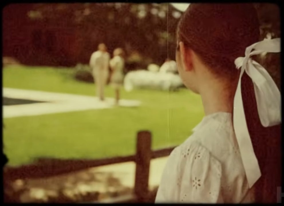 A person with a white ribbon in their hair looks toward two distant figures walking near a pool in a vintage-style setting