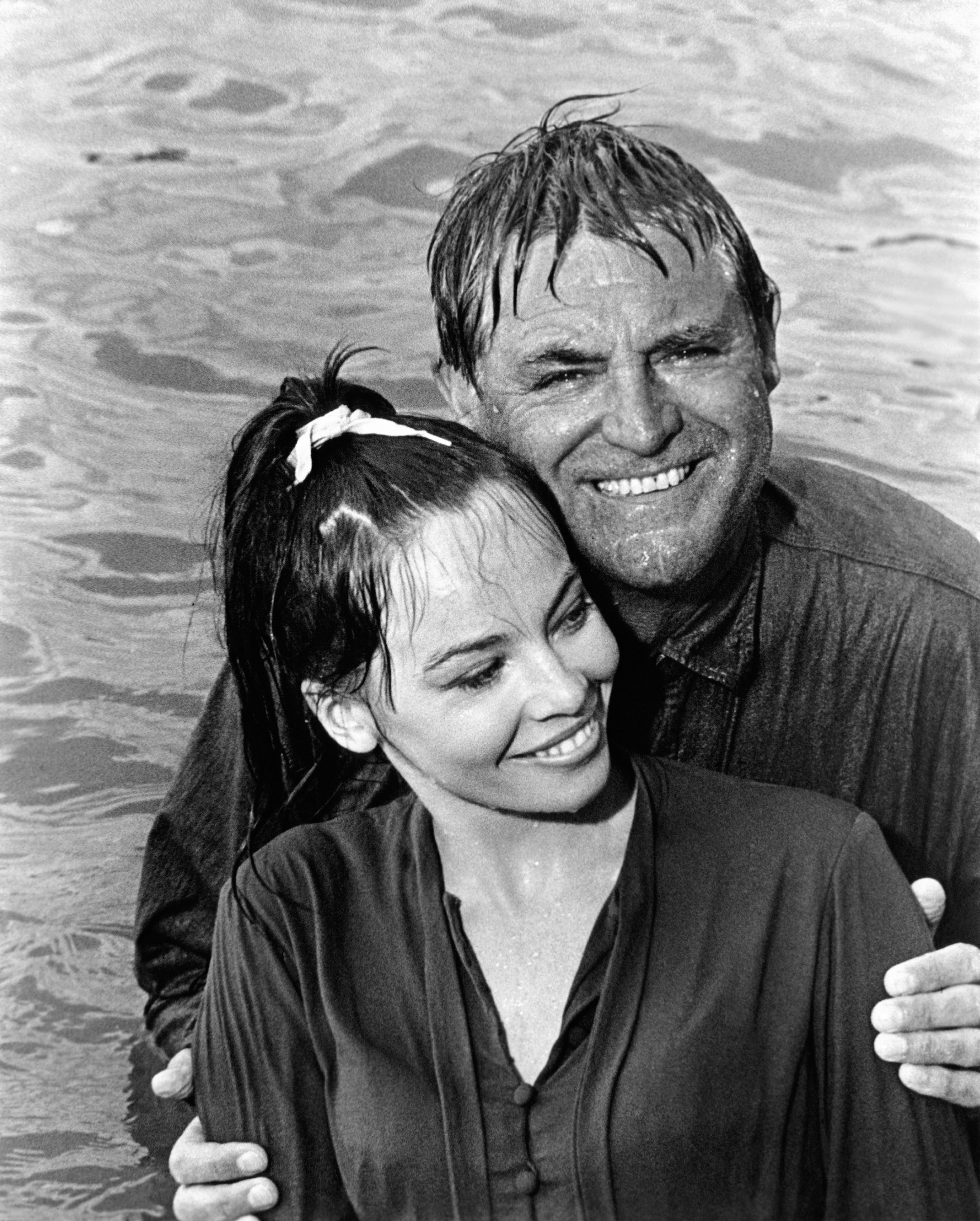 Two people smiling in a body of water, both wearing wet shirts. The man stands behind the woman, who has a ponytail