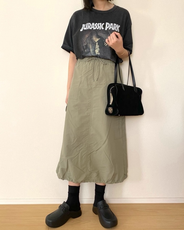Person in a &quot;Jurassic Park&quot; t-shirt, wearing a long skirt, black socks, clogs, and holding a black handbag