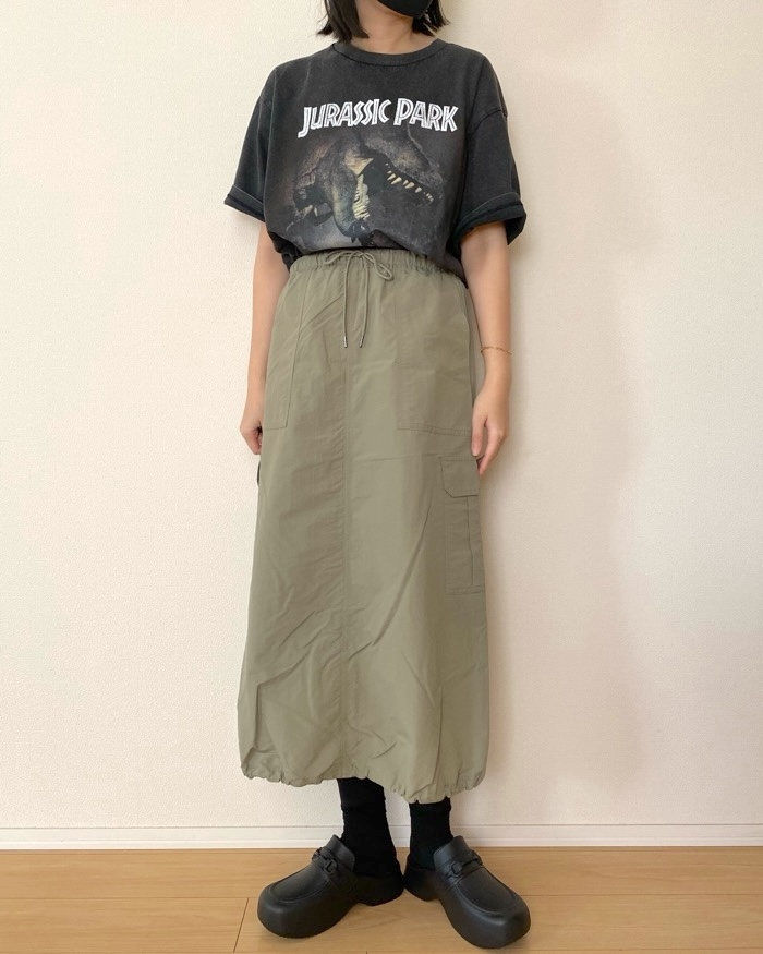 Person wearing a &quot;Jurassic Park&quot; T-shirt, long skirt, and clogs stands against a plain wall