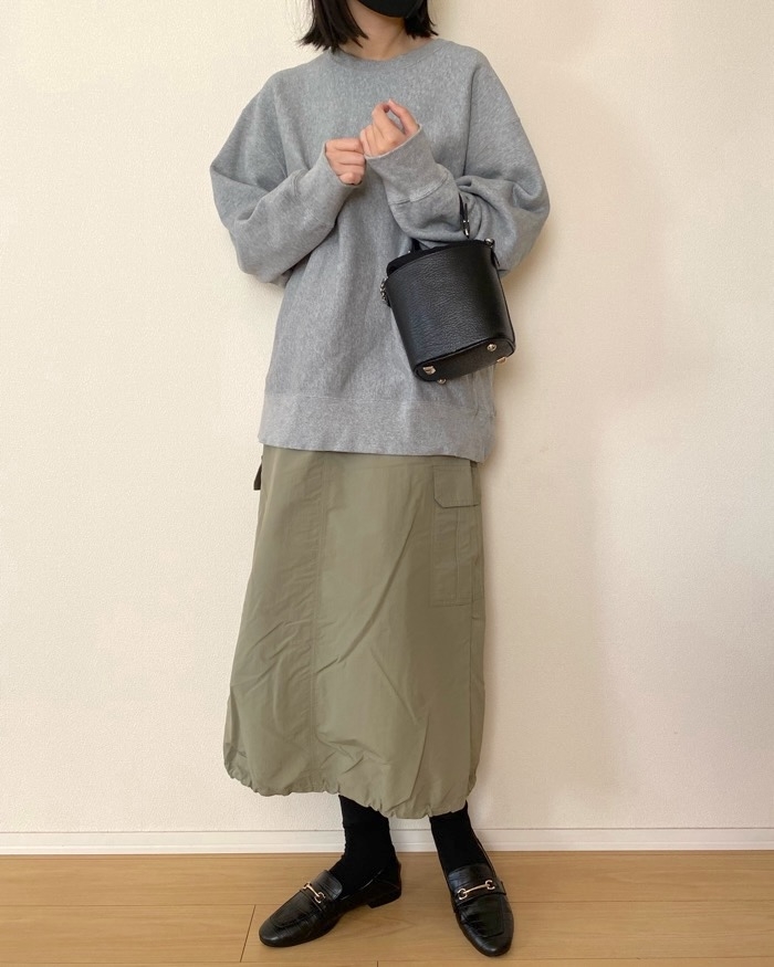 Person wearing an oversized sweater, long skirt, and loafers, holding a small handbag, standing indoors