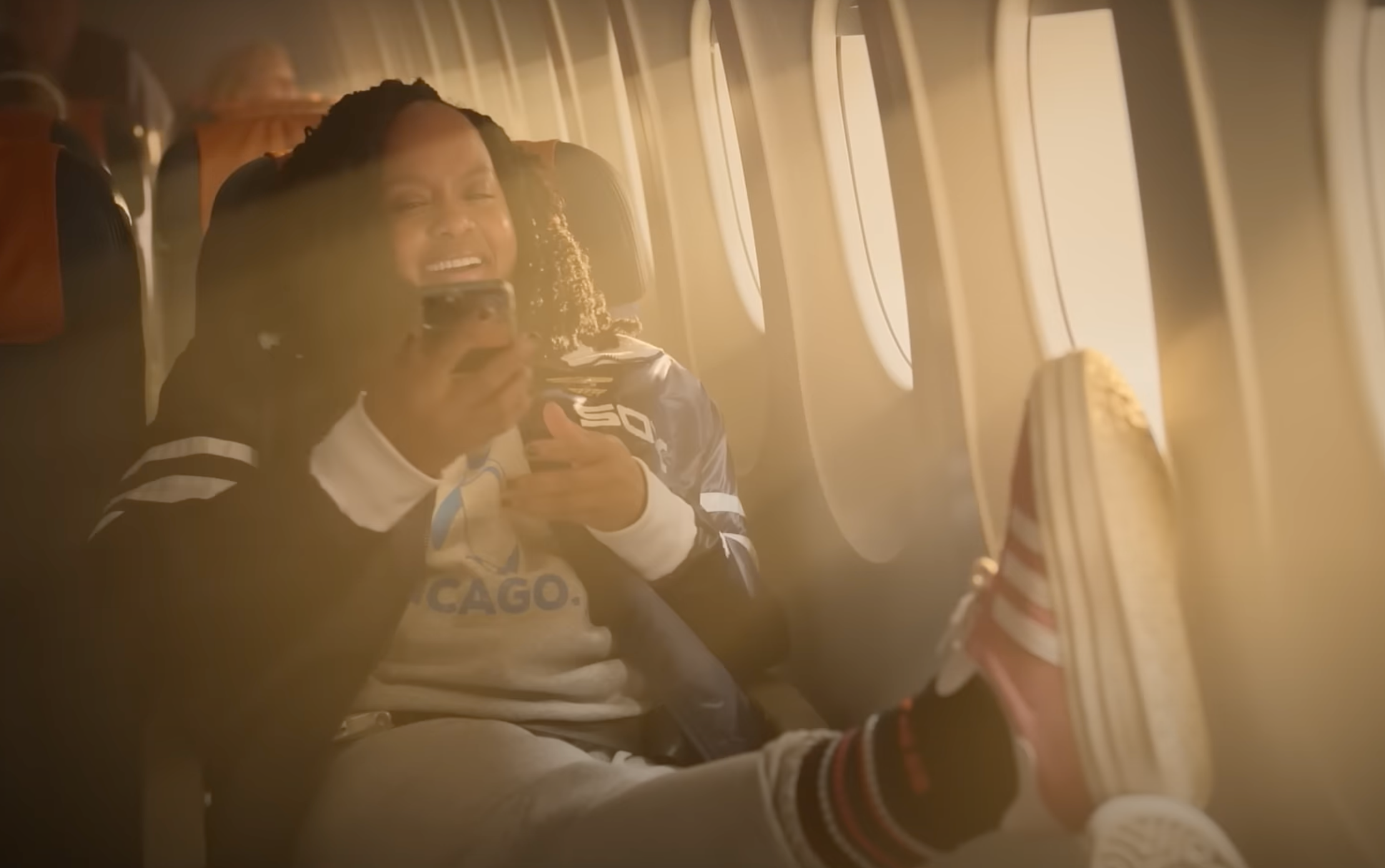 Person relaxing on an airplane seat, smiling while looking at a phone. Their foot is propped up on the window, suggesting a casual, comfortable vibe