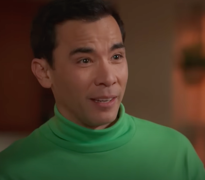 A person wearing a turtleneck shirt appears to be talking enthusiastically in a scene from a TV show or movie