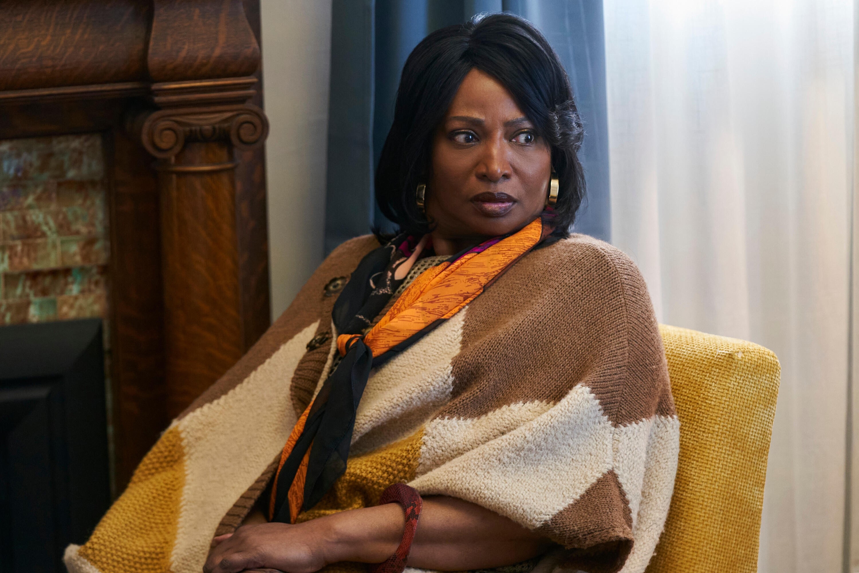 Person in a stylish geometric-patterned poncho sits, looking sideways with a thoughtful expression, in a setting resembling a movie or TV scene