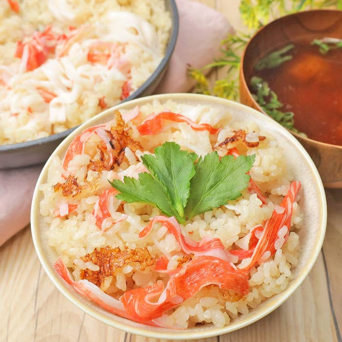 カニ風味の炊き込みご飯が盛られた茶碗。中央にパセリが飾られている。背景に同じ料理の鍋と味噌汁。