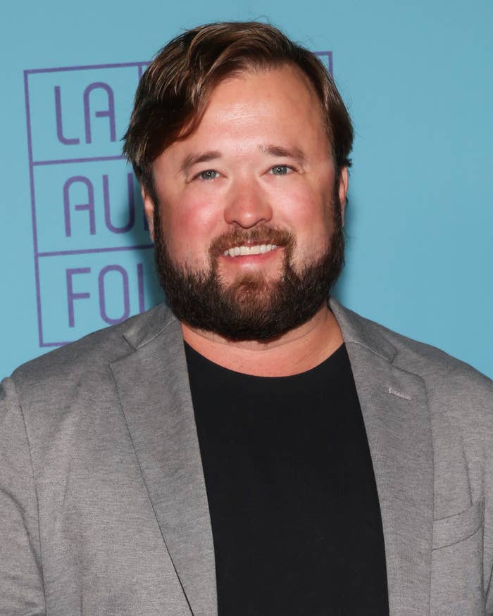 Haley Joel Osment smiling at an event, wearing a casual blazer over a black shirt, standing against a backdrop with text