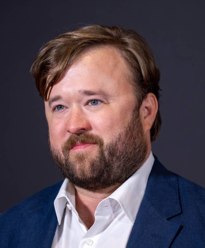 Haley Joel Osment with beard wearing a blue suit jacket and white shirt, looking slightly to the side