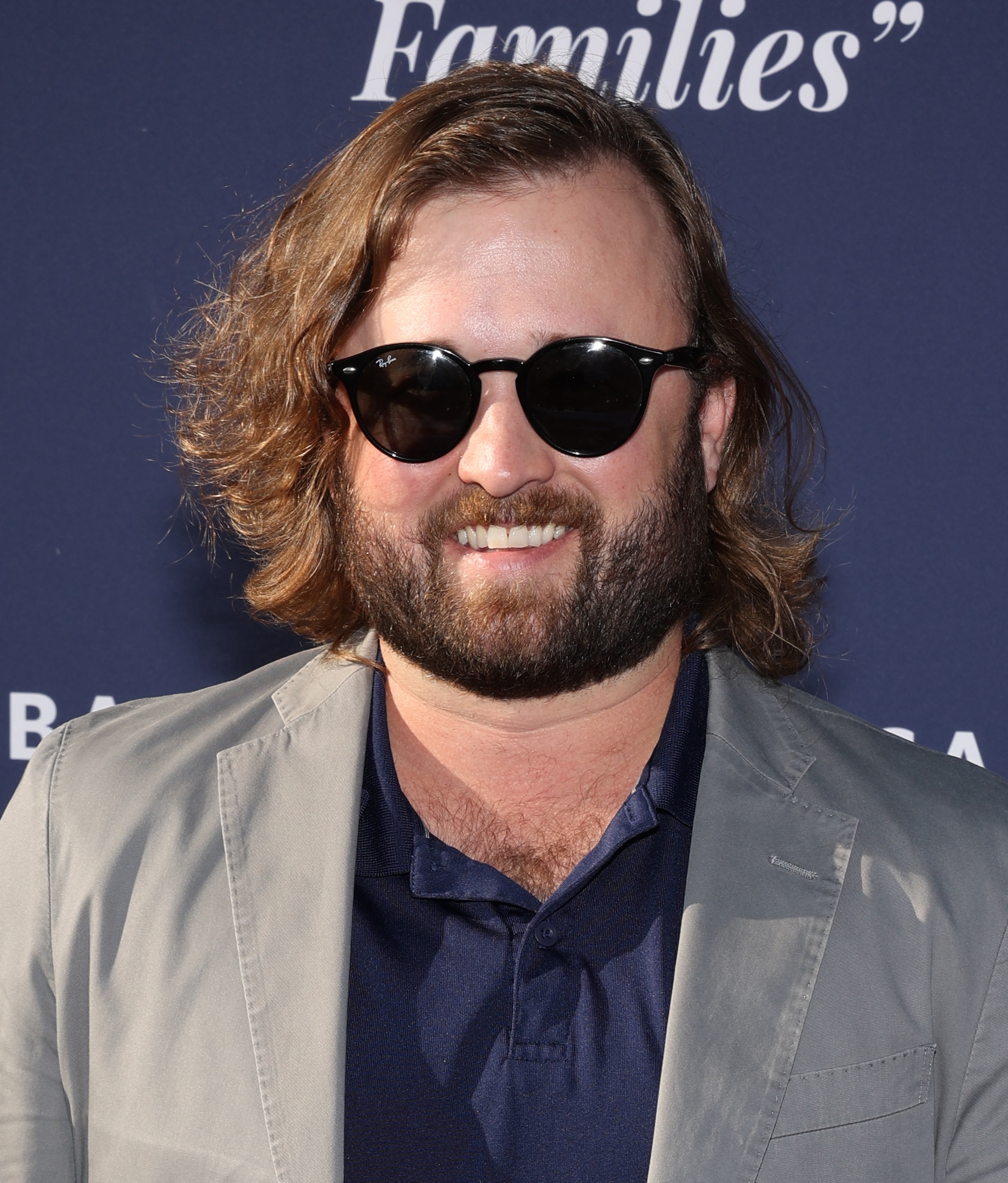 Haley Joel Osment with sunglasses and a beard smiles while wearing a casual suit and shirt at a formal event