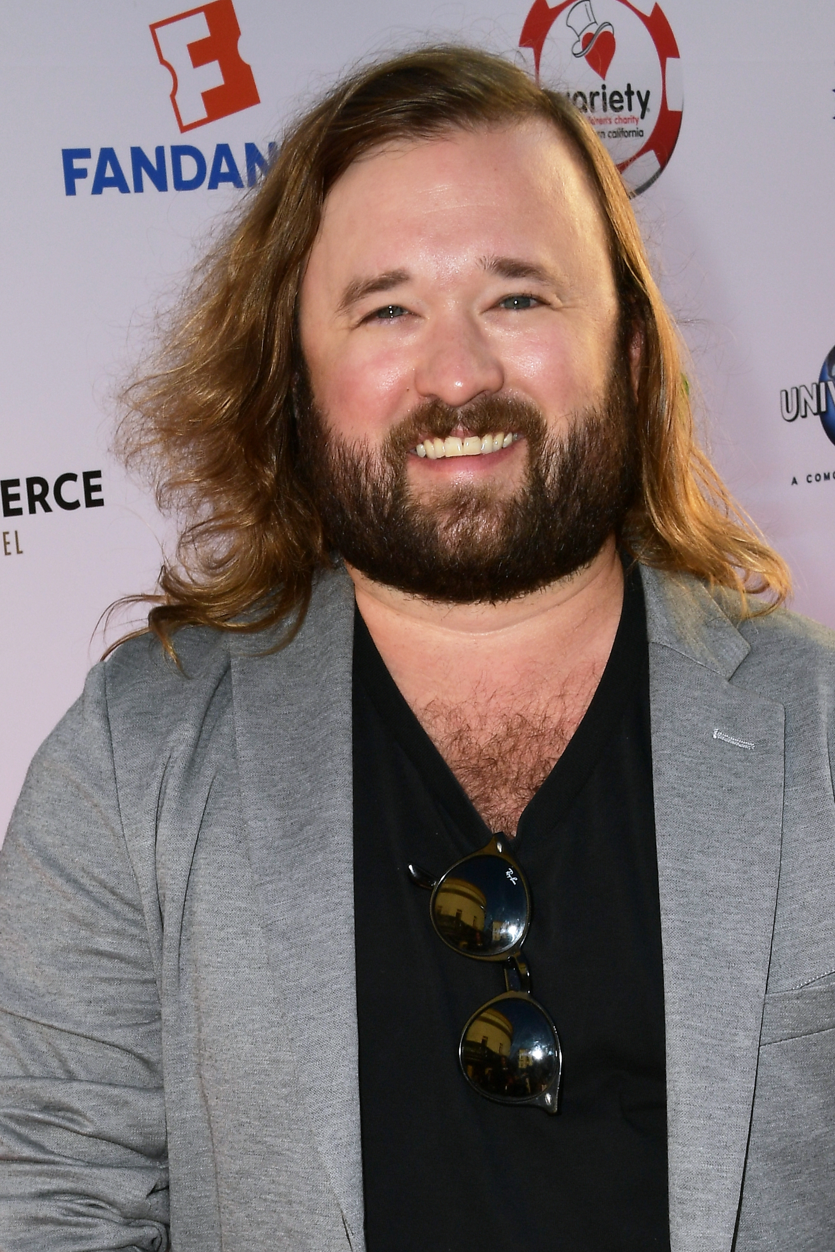 Haley Joel Osment with long hair and beard wearing a gray blazer over a black shirt, smiling at a media event backdrop