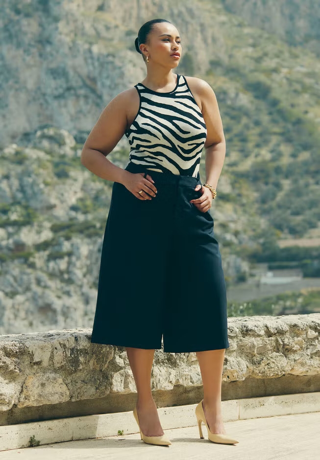 Person wearing a zebra-print top and wide-leg black pants poses confidently outdoors against a rocky landscape, wearing neutral heels