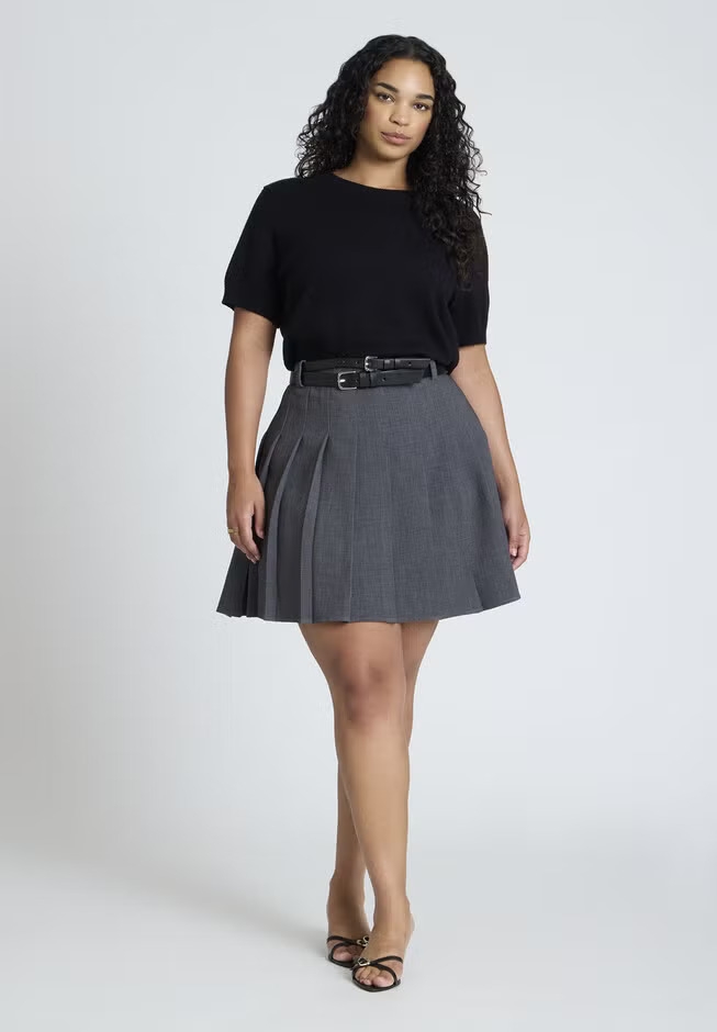 Person wearing a short-sleeve top, belted pleated skirt, and sandals, posing for a fashion photoshoot