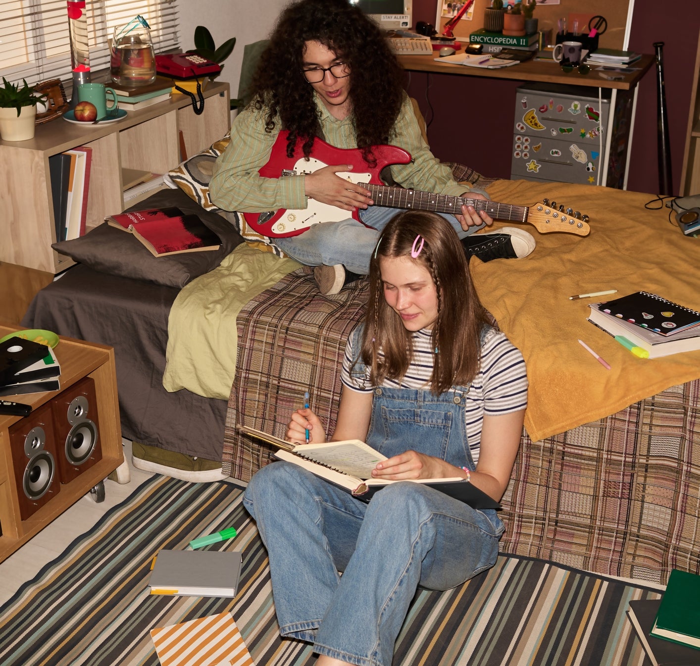 Two people are in a casual room; one is playing a guitar on the bed, the other is writing while sitting on the floor, surrounded by books