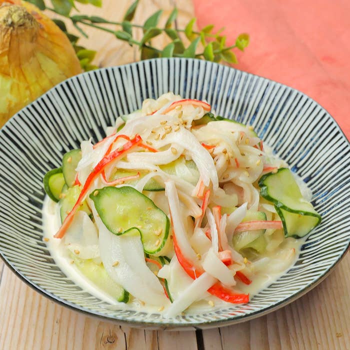 きゅうりとカニカマのサラダが皿に盛られ、ゴマがトッピングされています。背景に野菜が見えます。