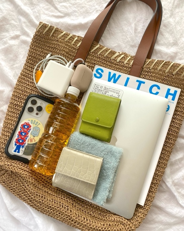 Woven bag containing a laptop, magazine, reusable bottle, phone with cartoon case, charging cables, and small accessories in neutral tones