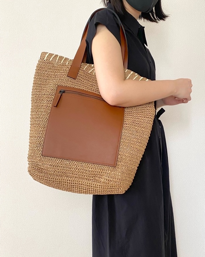 Person wearing a black dress holds a large straw tote bag with a front pocket and leather straps