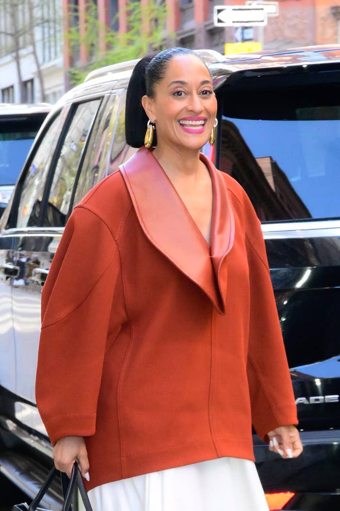 Tracee Ellis Ross smiling in a street, wearing a stylish oversized coat with a deep neckline and gold earrings. Ponytail hairstyle. Background of vehicles