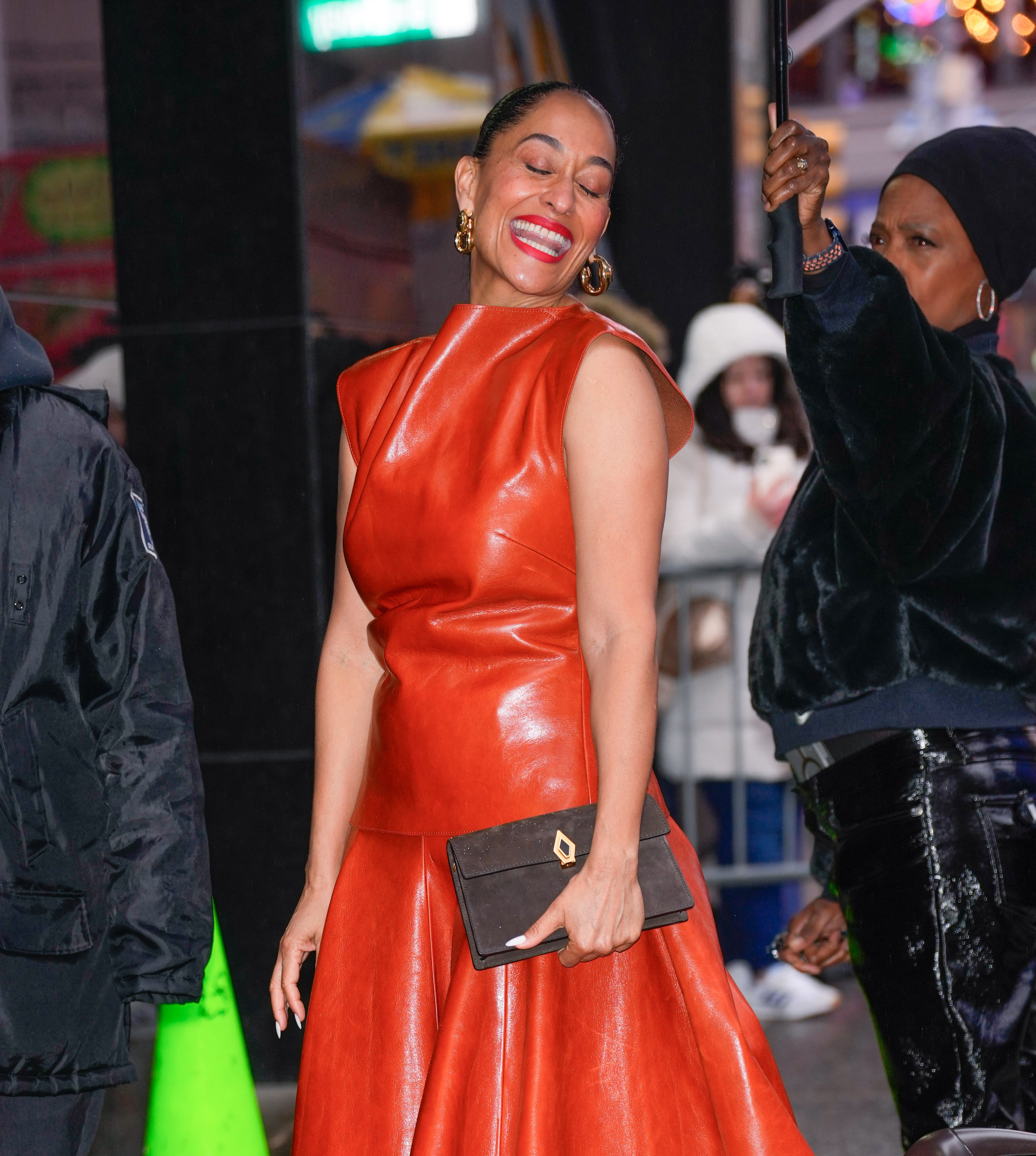 Tracee Ellis Ross smiling, wearing a sleek leather-like dress and holding a clutch, on a city street. Another person adjusts something nearby
