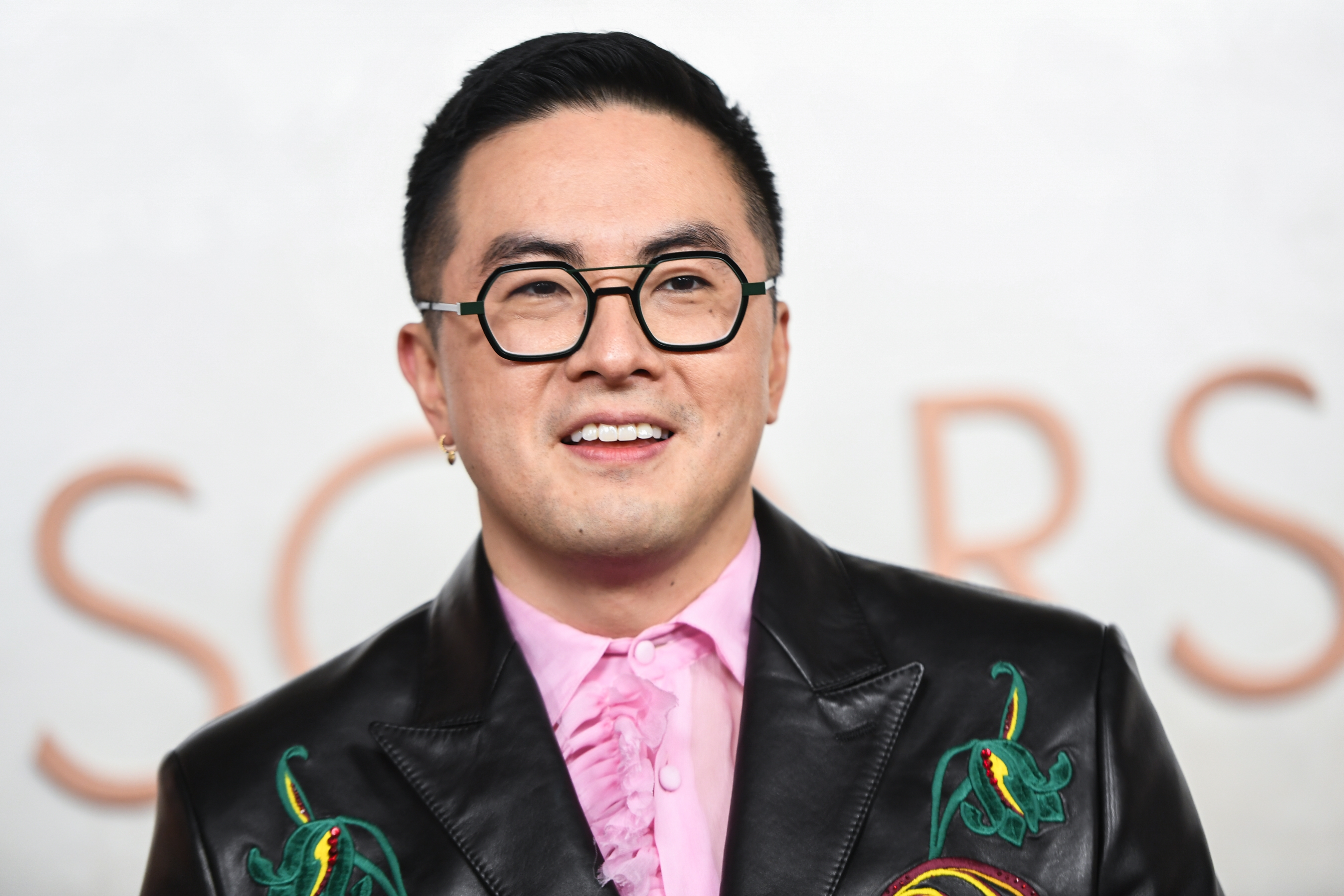 Bowen Yang in a black leather jacket with floral embroidery and a pink ruffled shirt, smiling at an event