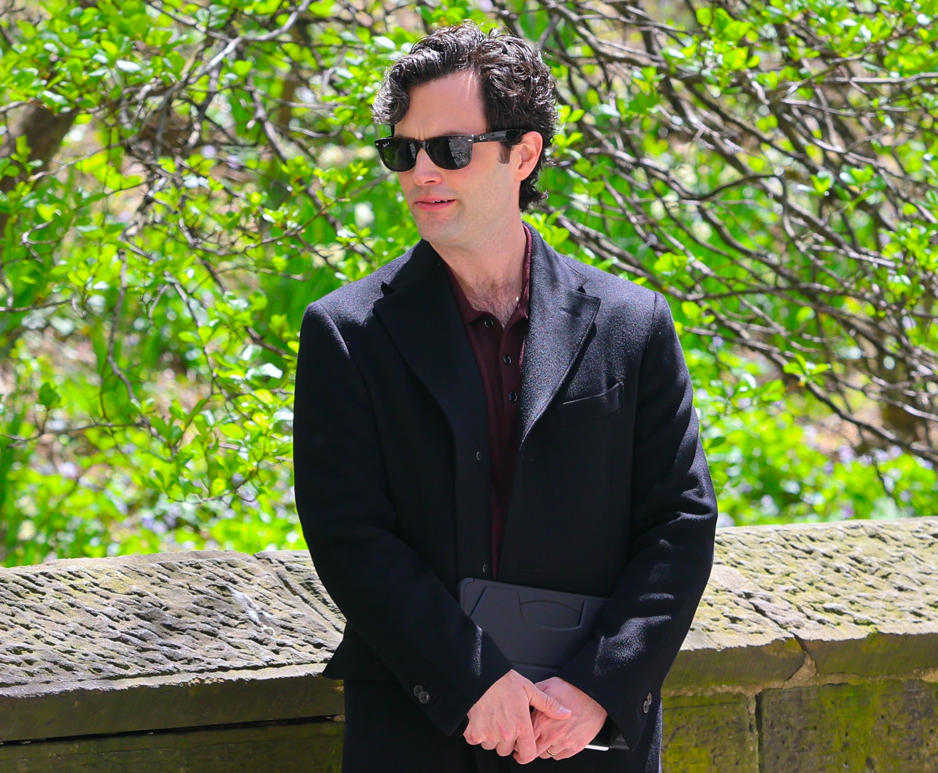 Penn Badgley with sunglasses and dark coat stands outdoors against a stone wall, with green foliage in the background