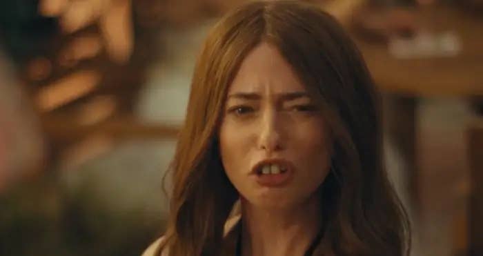 Aimee Lou Wood with long brown hair has an open-mouthed expression, appearing to be talking or singing, against a blurred background