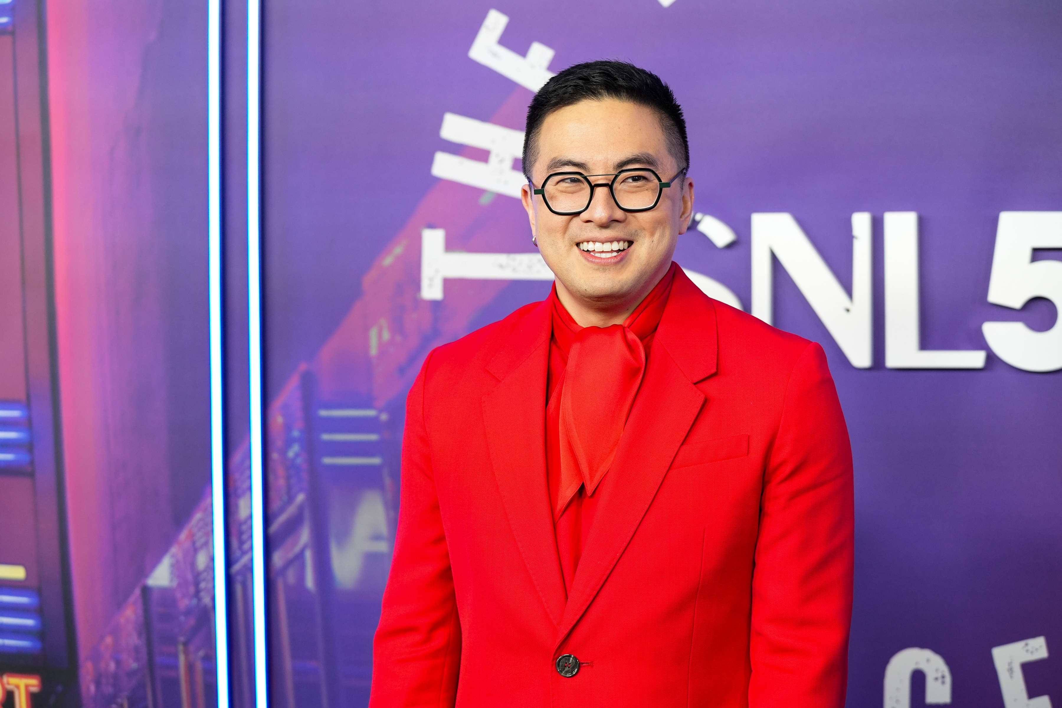 Bowen Yang in a stylish, tailored suit with a matching scarf, standing in front of a backdrop with "SNL" visible