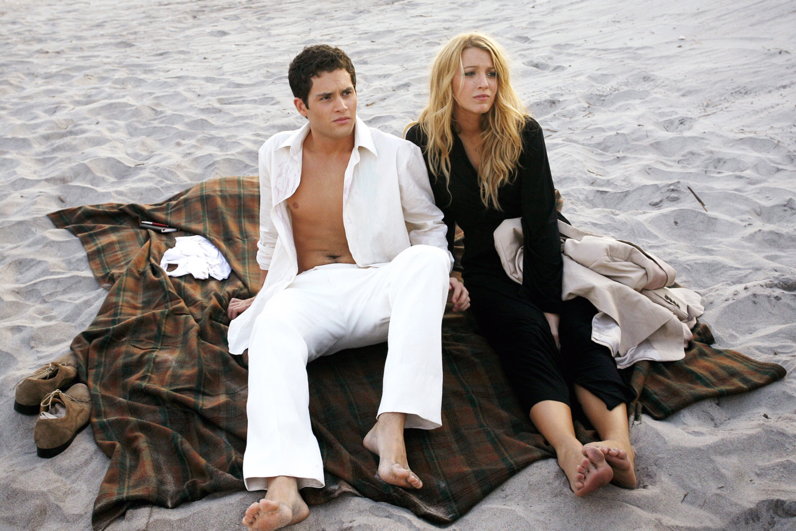 Penn Badgley and Blake Lively sitting on a plaid blanket on the beach, dressed in casual, light clothing, looking away from the camera