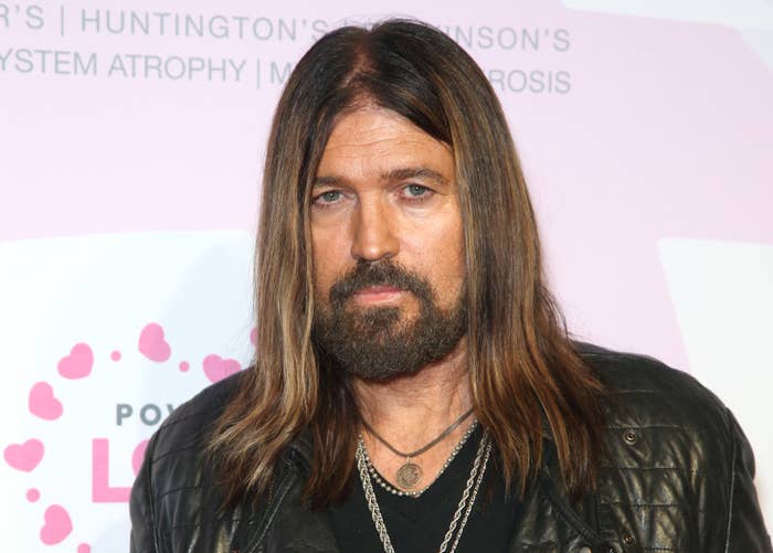 Billy Ray Cyrus with long hair and beard wearing a leather jacket and layered necklaces at an event with a backdrop featuring heart patterns