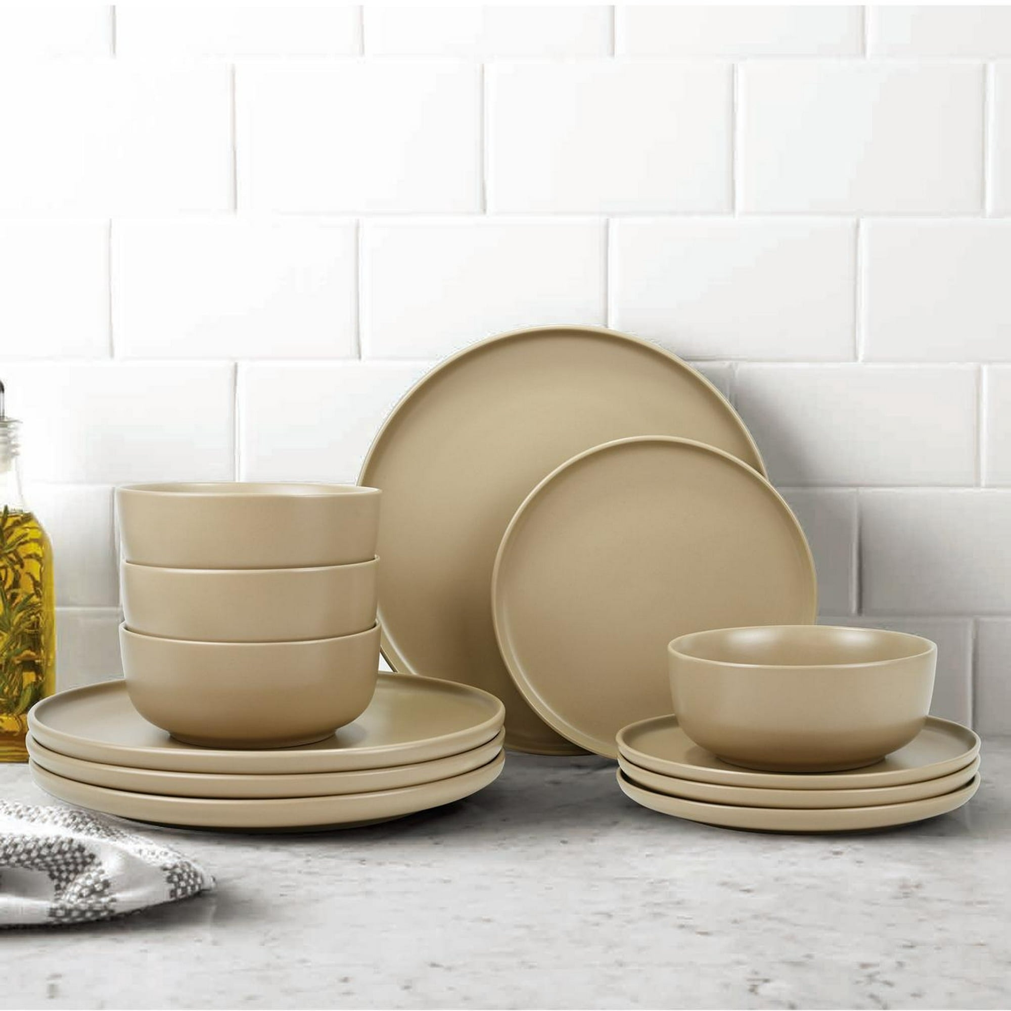 Minimalist beige dinnerware set with plates and bowls displayed on a kitchen counter in front of a tiled backsplash. White towel visible