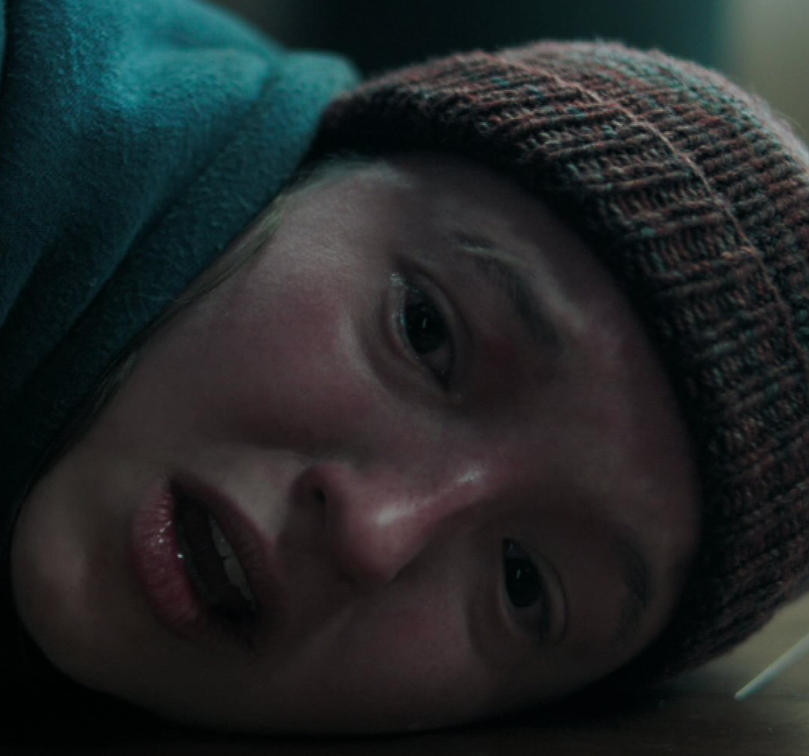 Persona con gorro de punto mira preocupada hacia adelante, aparentemente acostada en el suelo. Escena transmitiendo tensión o angustia