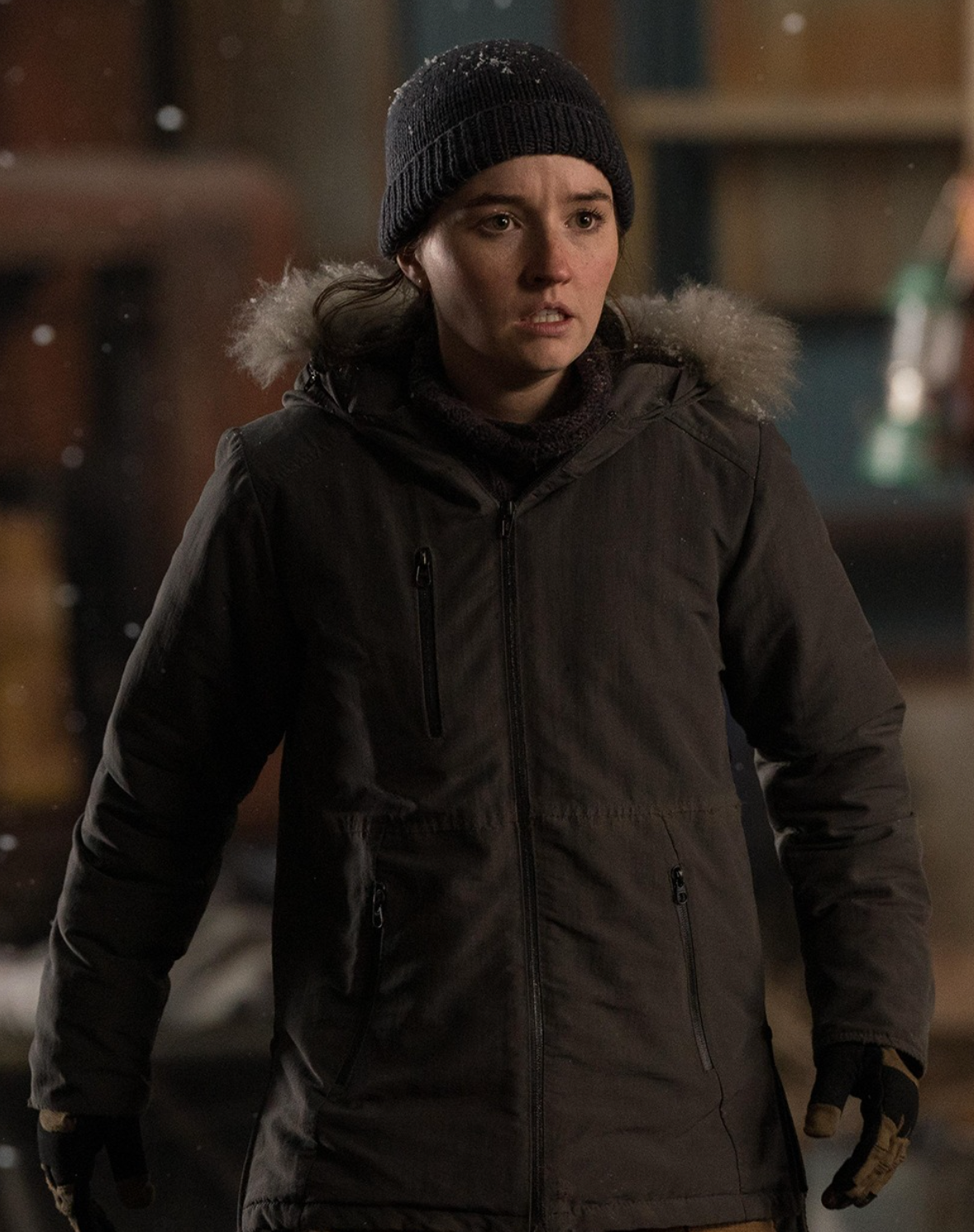 Persona con abrigo y gorro de invierno, parece estar en una escena de suspenso. Nieve cae a su alrededor, sugiriendo un entorno frío