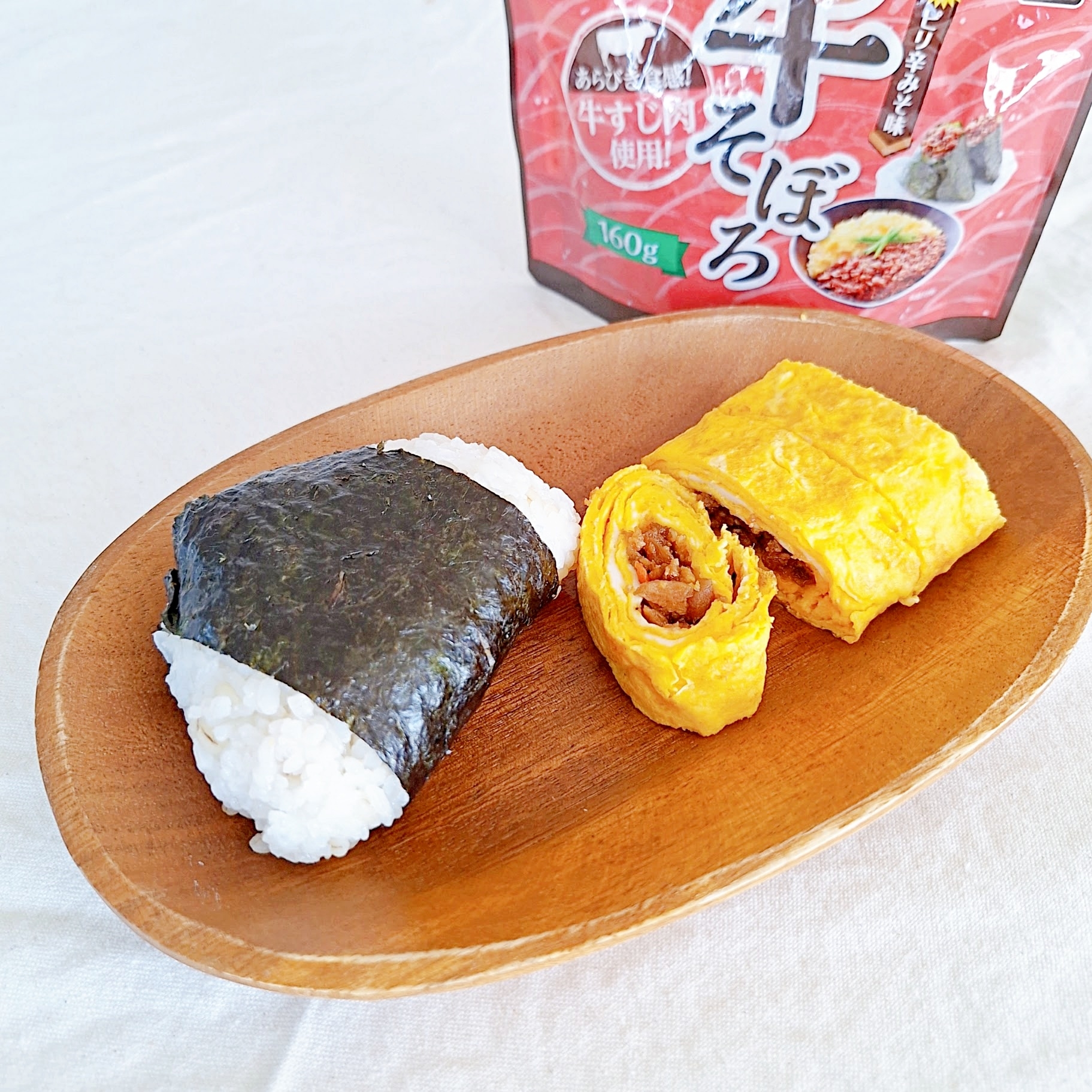 木の皿に海苔巻きおにぎりと牛肉入り卵焼きが載っている。背景に食品パッケージが見える。