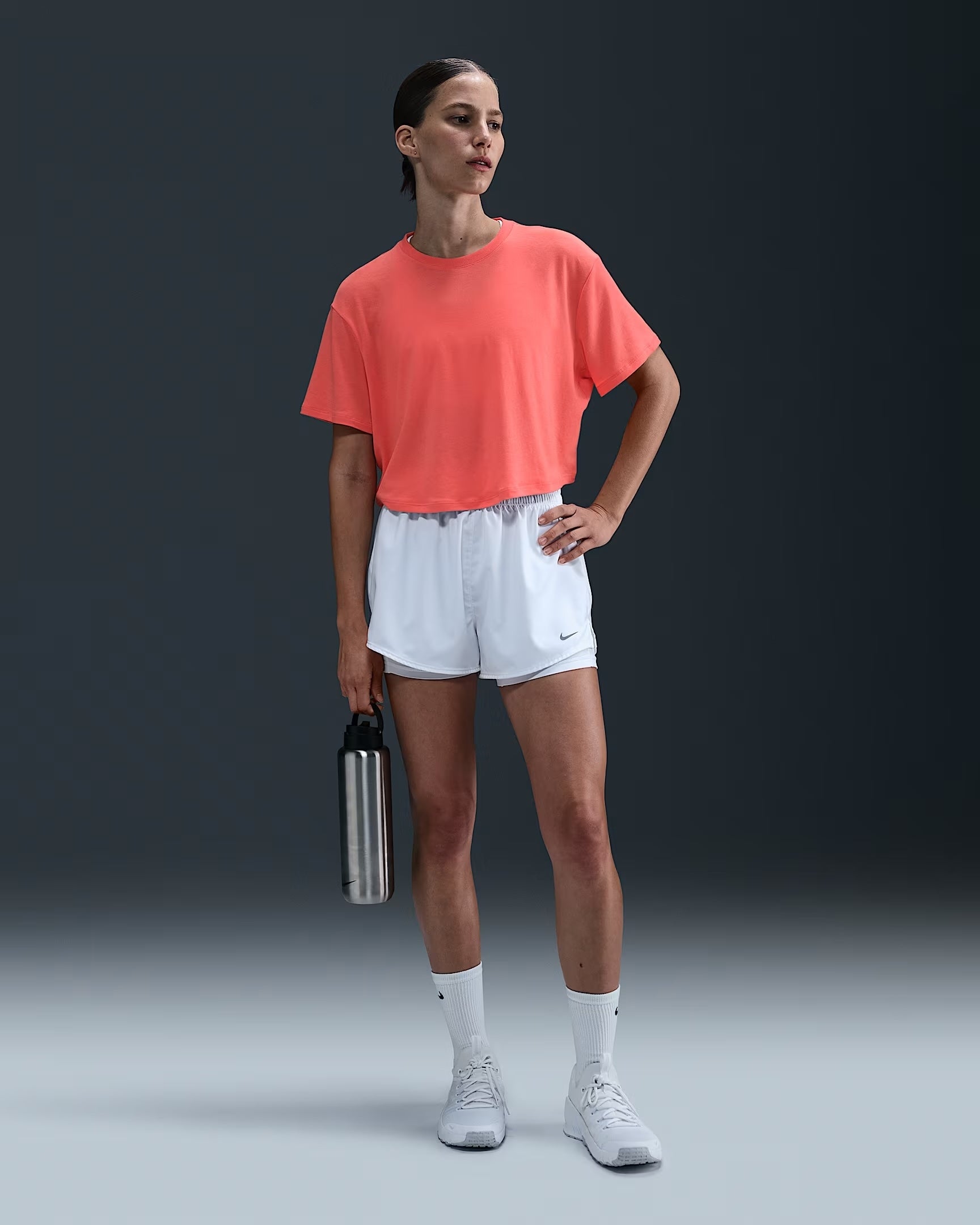 Seorang wanita berpose dalam pakaian sporty kasual, mengenakan atasan longgar, celana pendek, dan sepatu kets, memegang botol air logam