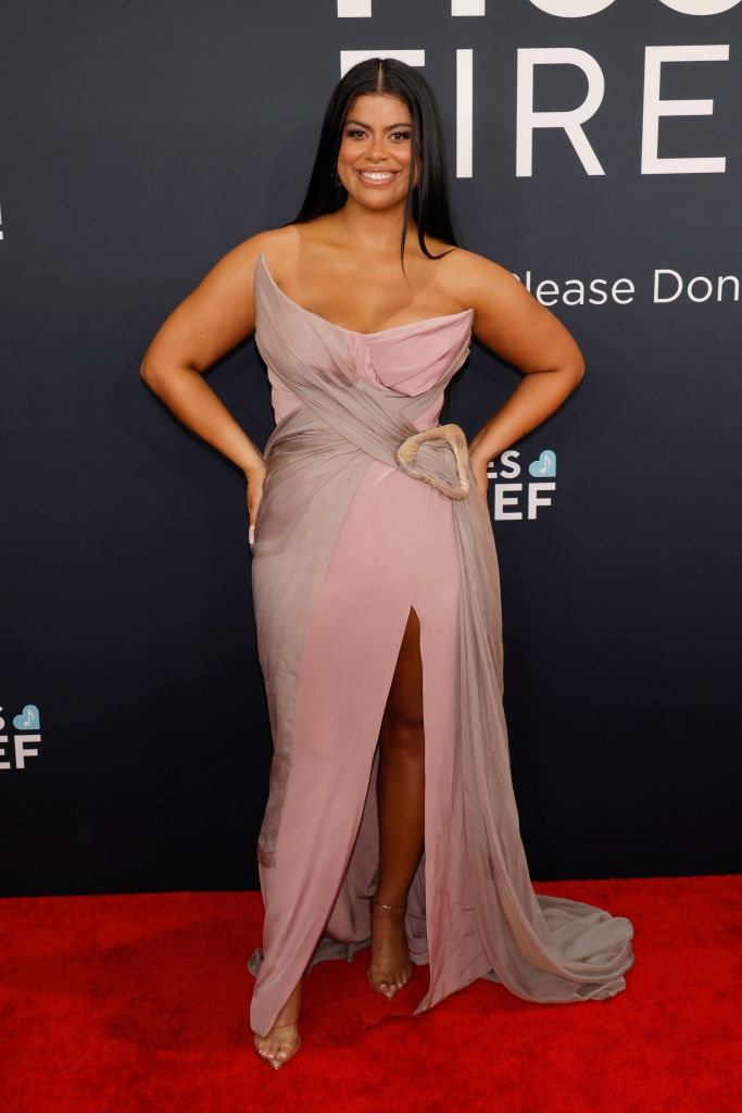 Person in elegant, strapless gown with a gathered waist and thigh-high slit at a red carpet event, posing confidently with hands on hips