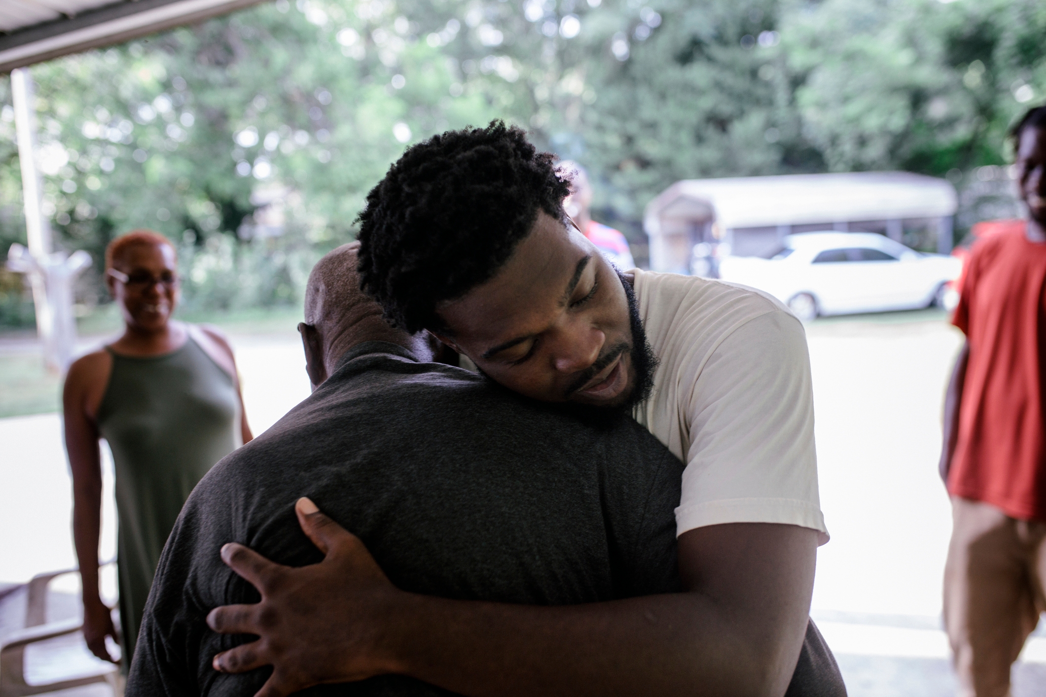 Two people share a heartfelt embrace outdoors, with others nearby, conveying a sense of family and connection