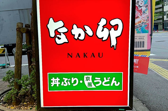 Street sign for Nakau restaurant, promoting rice bowl and Kyoto-style udon. Colorful signs and a sidewalk are visible in the background