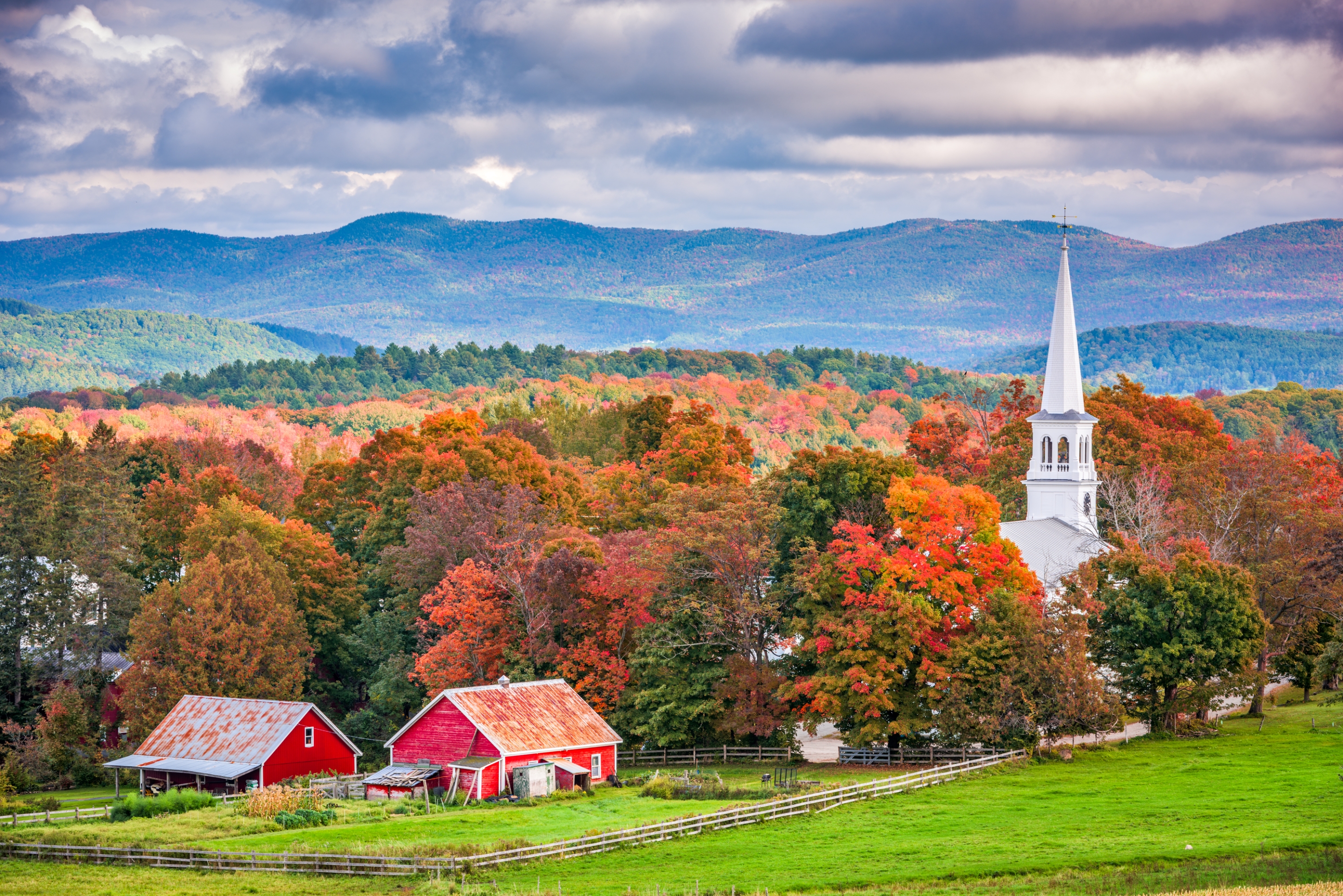 Scenic rural landscape with red barns, a white steeple church, and rolling hills under a cloudy sky. Autumn foliage colors the trees