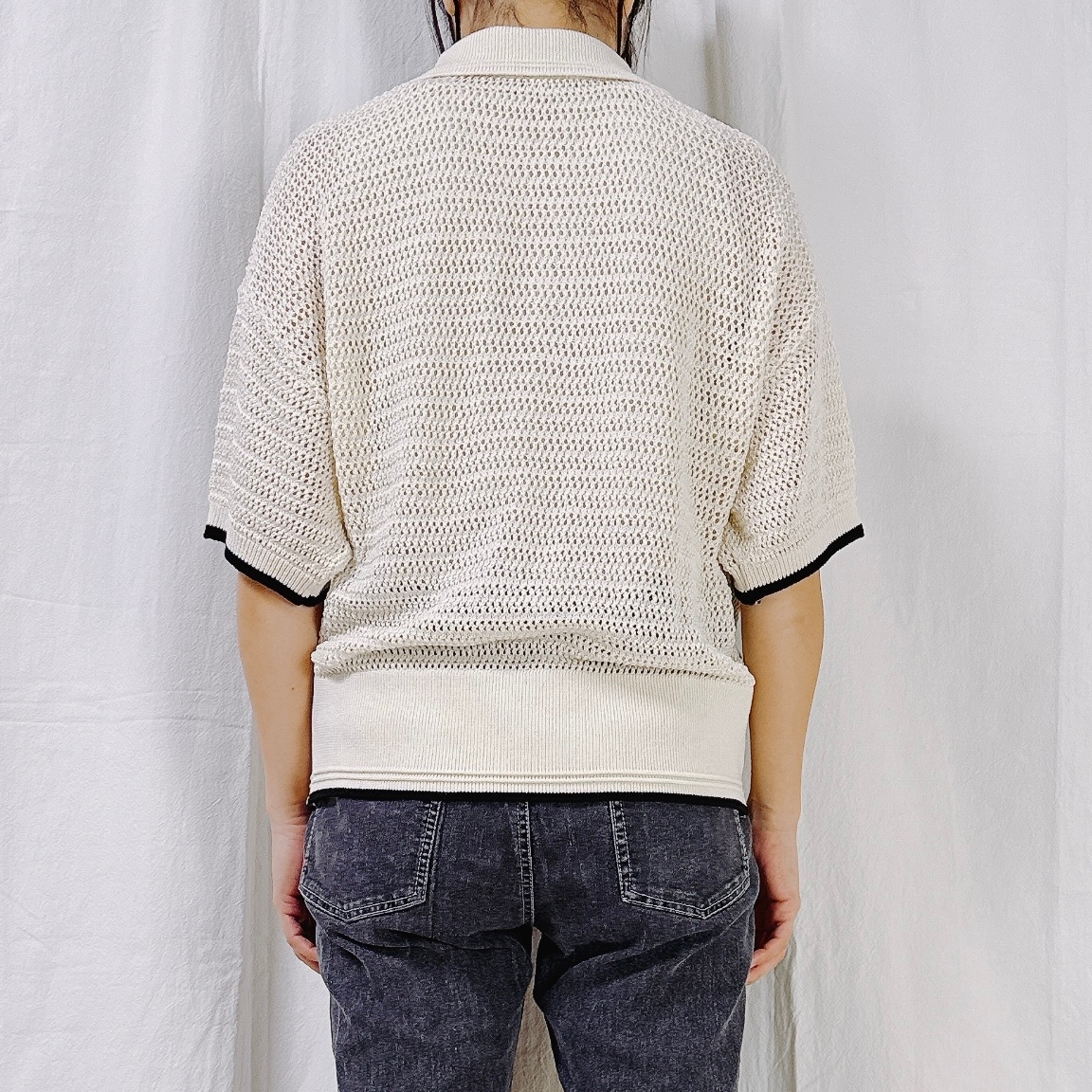 Person facing away, wearing a light knit top and dark jeans, standing against a plain backdrop