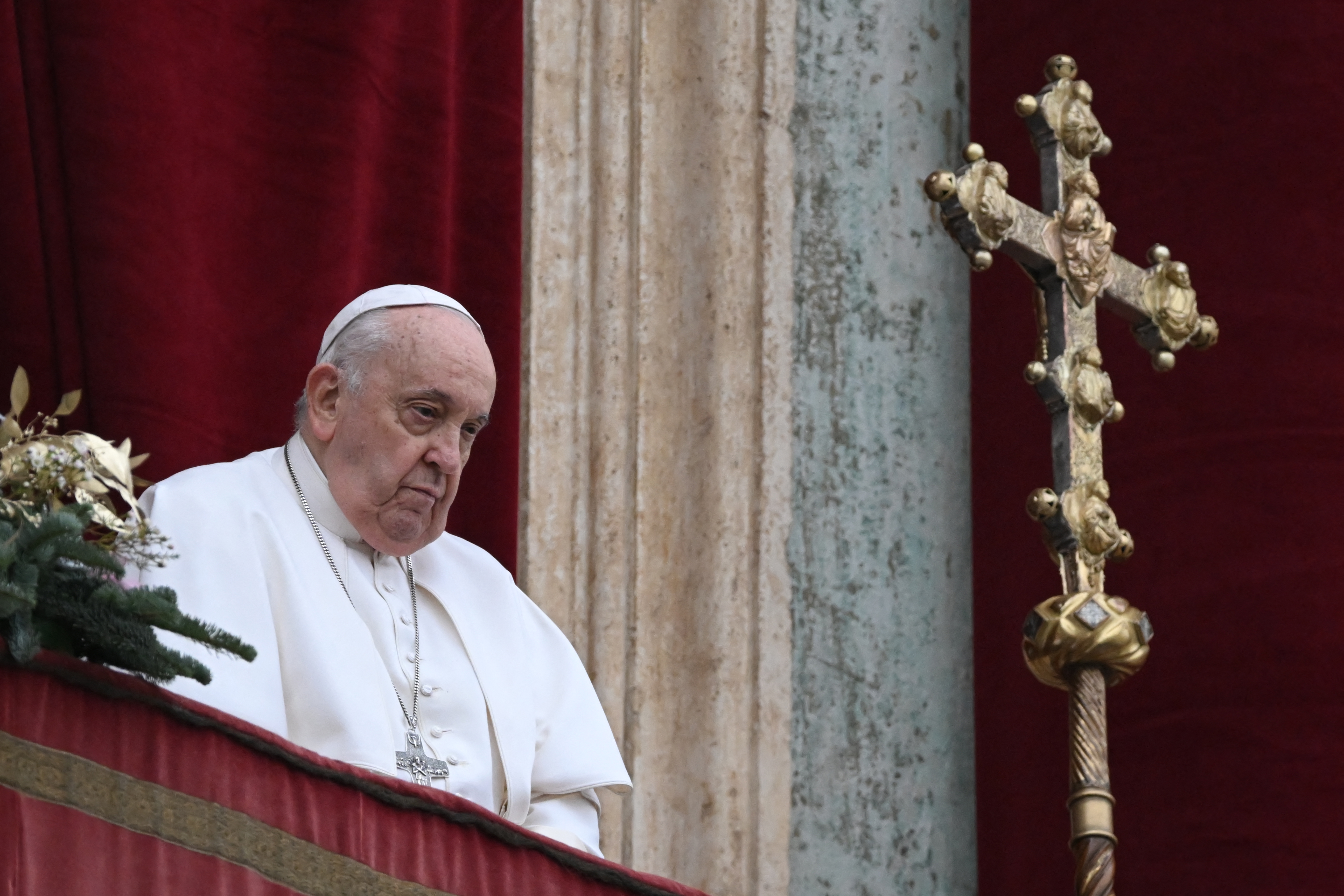 A religious leader in white attire stands on a balcony beside an ornate golden cross