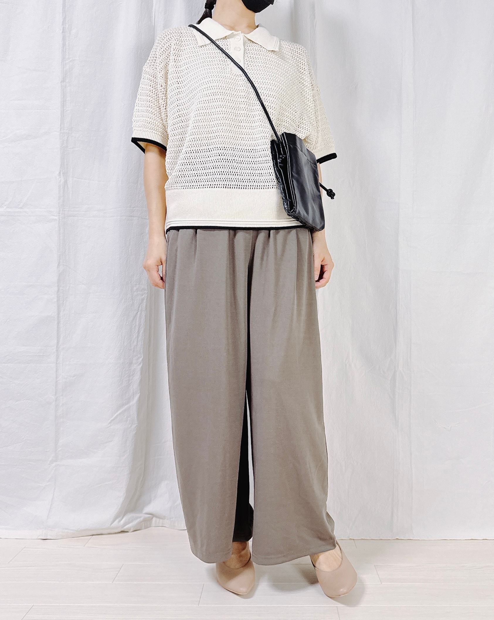 Person wearing a loose-knit short-sleeve top, wide-legged trousers, crossbody bag, and flats, posing against a plain backdrop