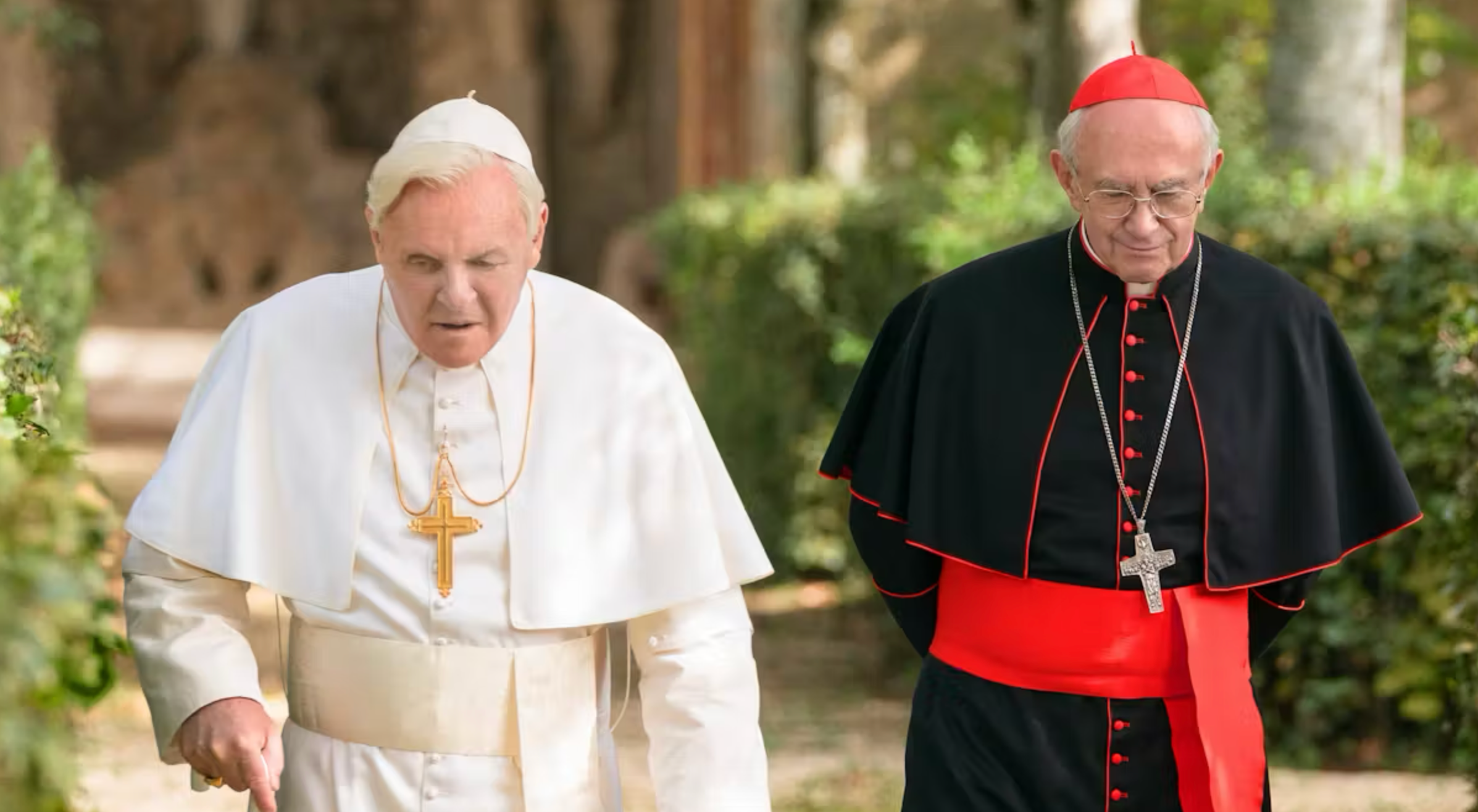 Dos hombres mayores en atuendos religiosos, uno con ropa blanca y otro con negra y roja, caminan juntos al aire libre