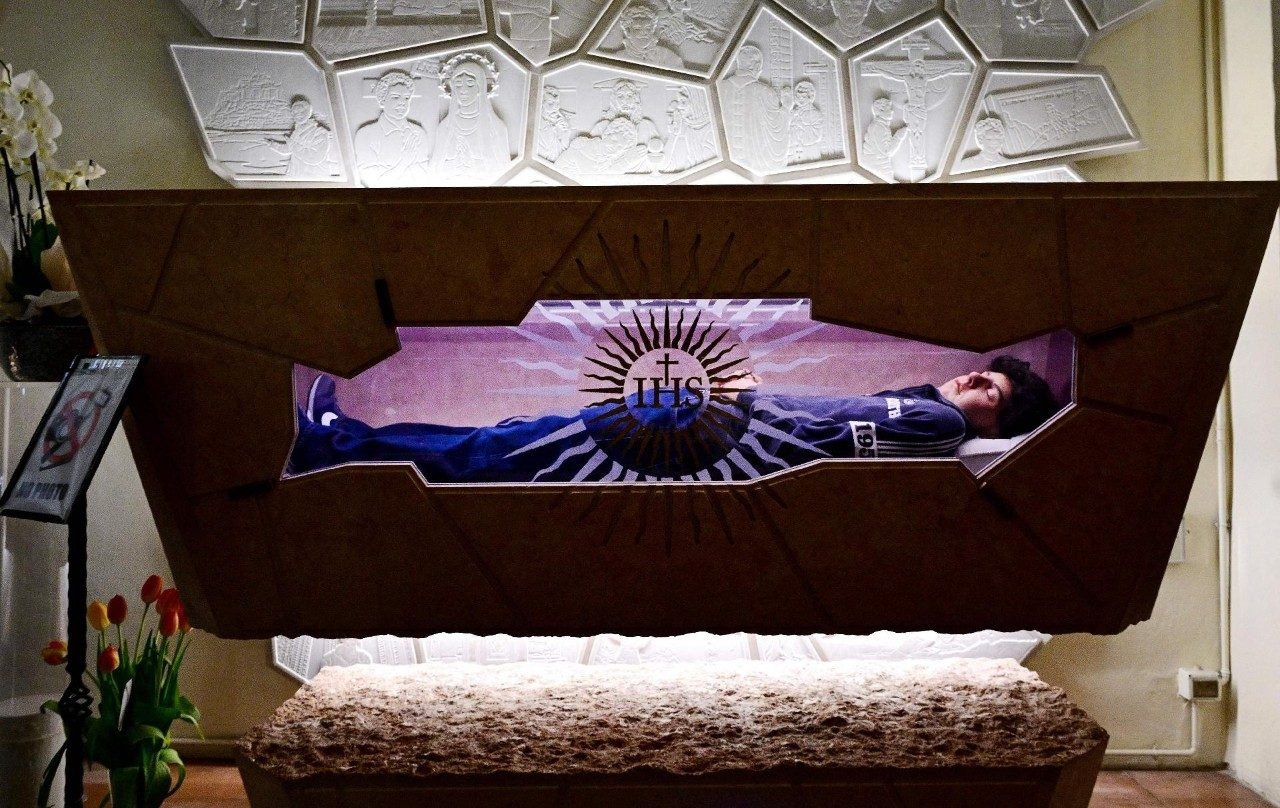 Una figura yace en reposo dentro de una urna iluminada, en un entorno decorativo con relieves artísticos