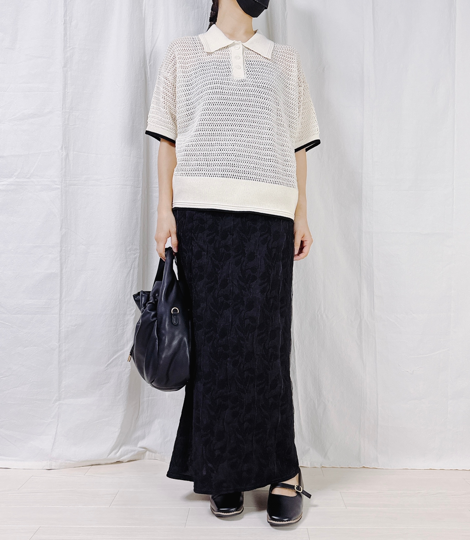 Person in a casual outfit with a knitted short-sleeve top, long skirt, holding a bag, standing against a plain backdrop. Faces are not visible