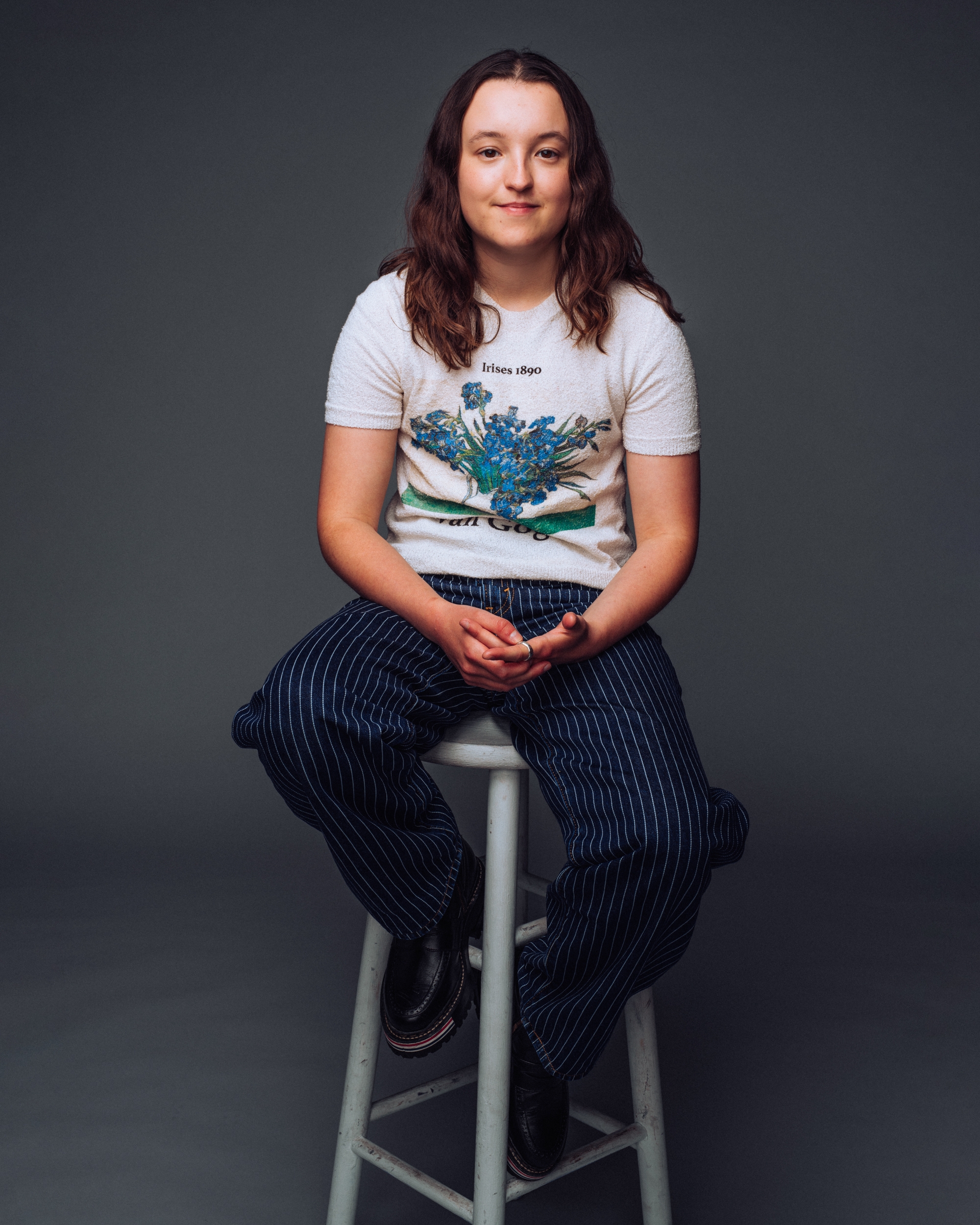 Bella Ramsey seated, wearing a graphic t-shirt with floral design and pinstripe pants