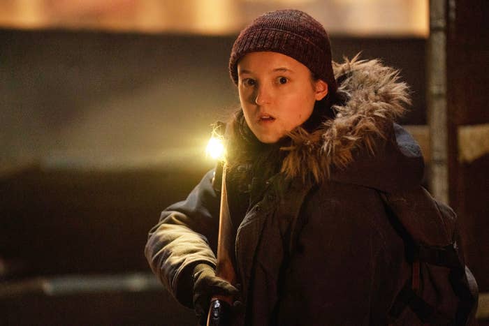 Bella Ramsey as Ellie in a winter coat and beanie holds a lit torch, looking surprised in a dimly lit setting
