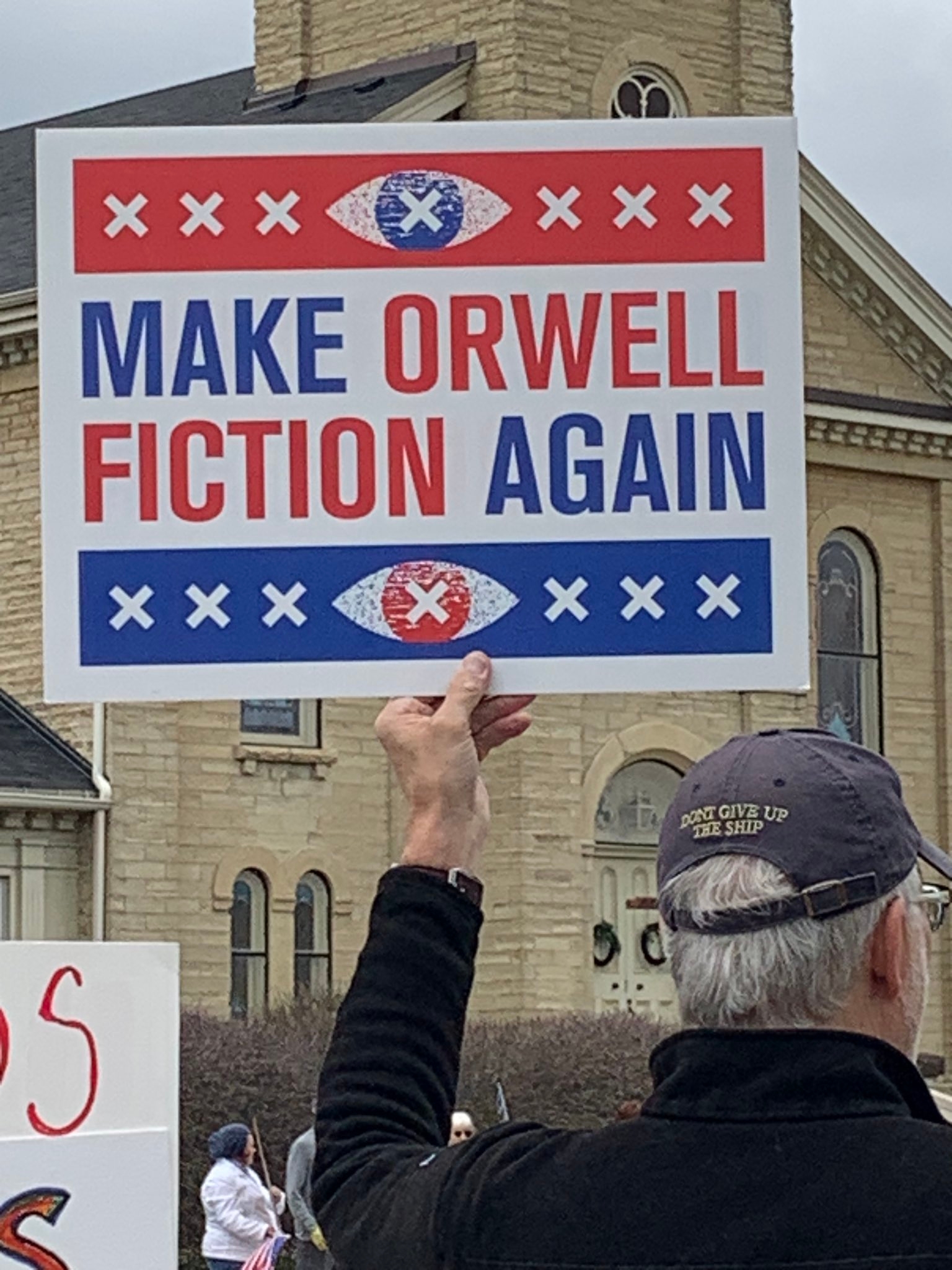 Person holds a protest sign reading, "Make Orwell Fiction Again," referencing George Orwell's dystopian themes