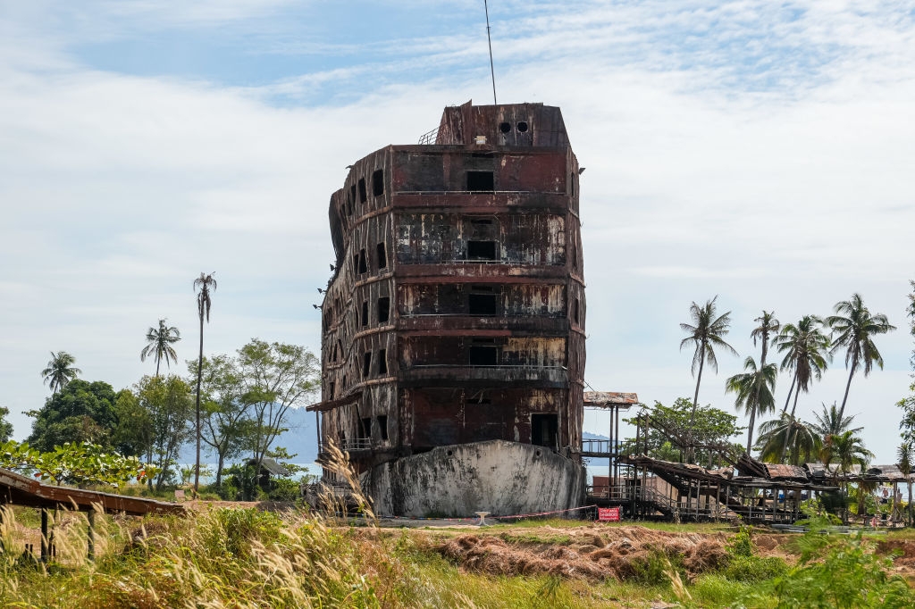 Abandoned, burned multi-story building stands in a field surrounded by palm trees under an overcast sky