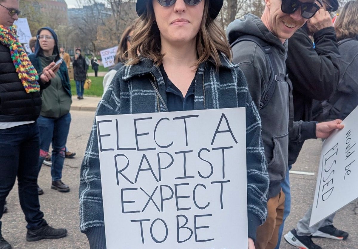 Person at a protest holding a sign that reads, &quot;ELECT A RAPIST EXPECT TO BE FUCKED
