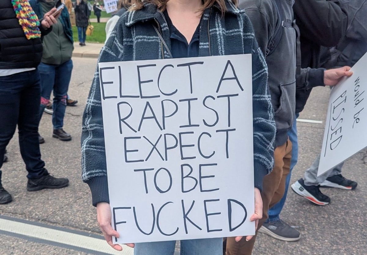 Person at a protest holding a sign that reads, "ELECT A RAPIST EXPECT TO BE FUCKED