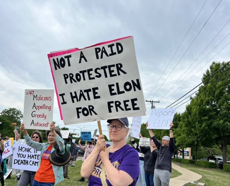 Person holding sign saying &quot;Not a paid protester. I hate Elon for free,&quot; with a group of protesters holding various signs in the background