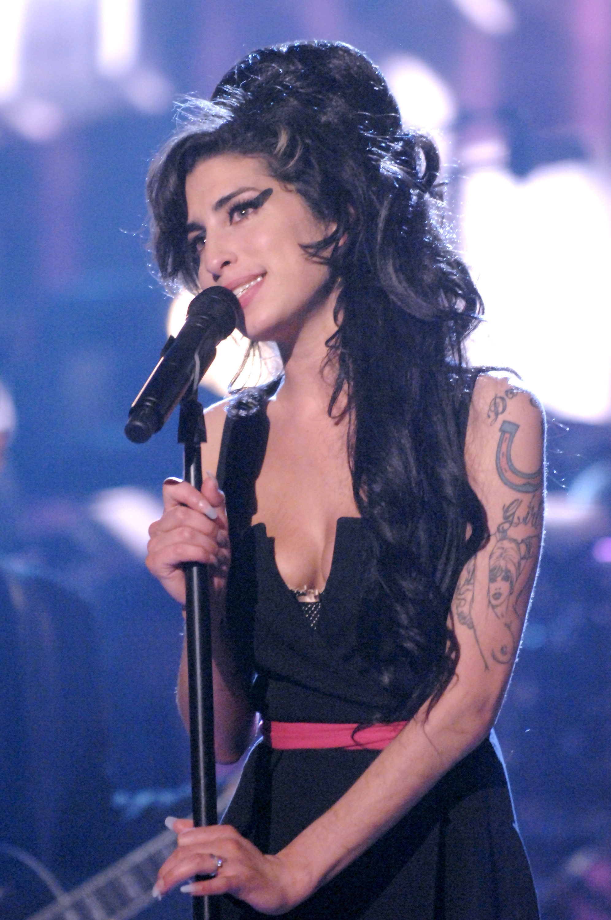 Singer with a beehive hairstyle performs on stage, wearing a black dress with a plunging neckline and tattooed arms